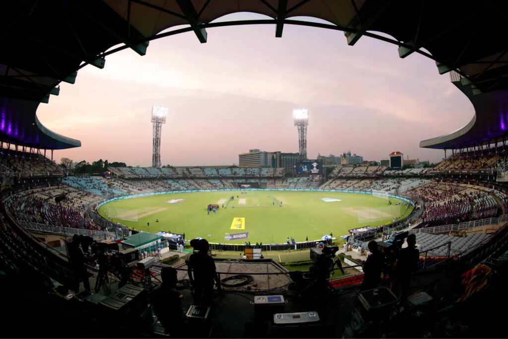 Wide-angle view of a packed cricket stadium at sunset, with a green field and illuminated stands.