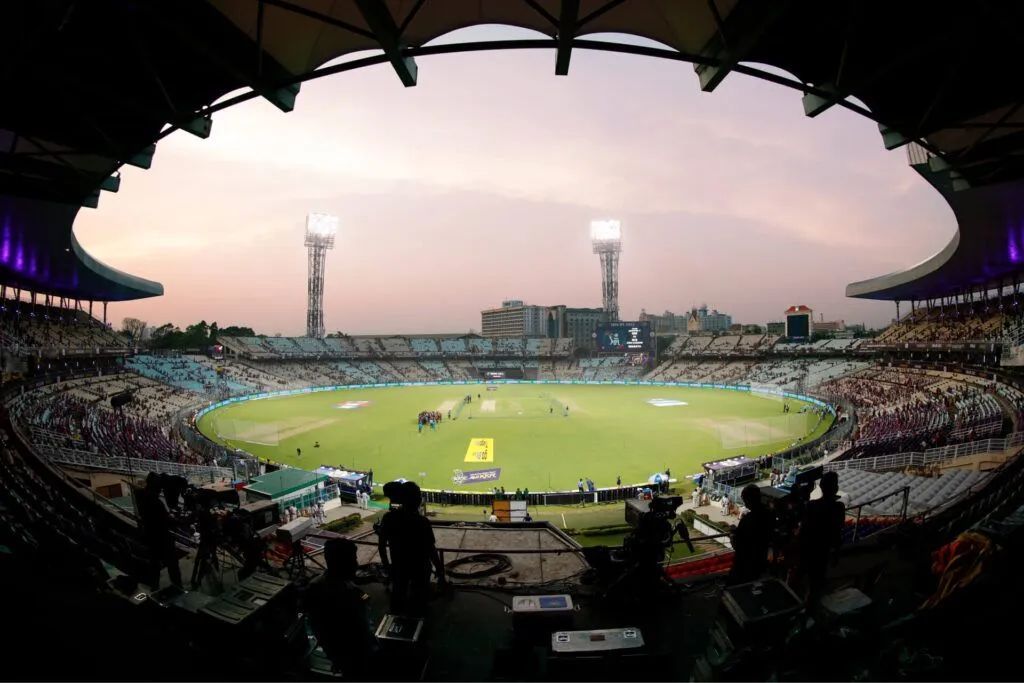 Cricket stadium packed with fans at dusk, green field lit under a curved roof.