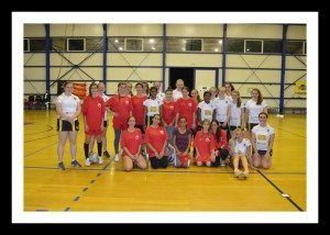 Group of volleyball players posing indoors on a gym court, many in red uniforms with a net behind them