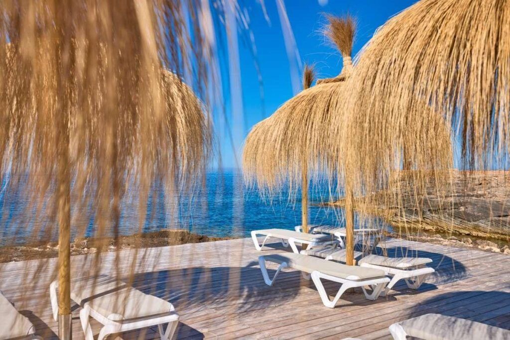 Beach loungers under thatched umbrellas by the sea, with blue water and clear sky