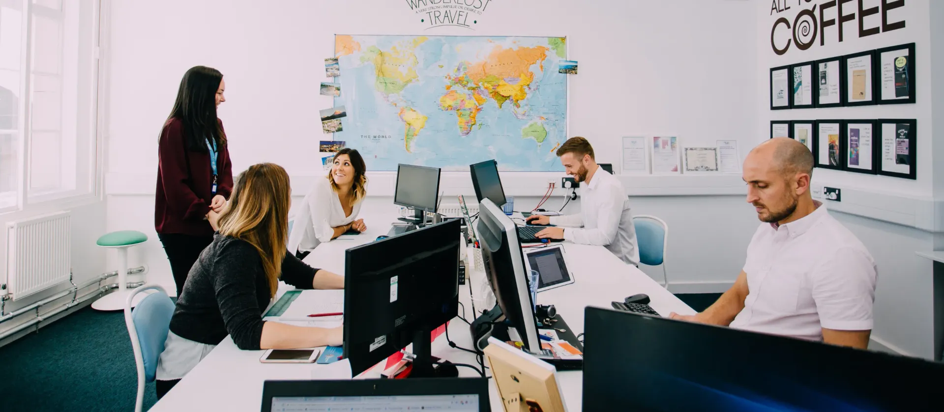 Office meeting with four people around desks, whiteboard and world map in the background