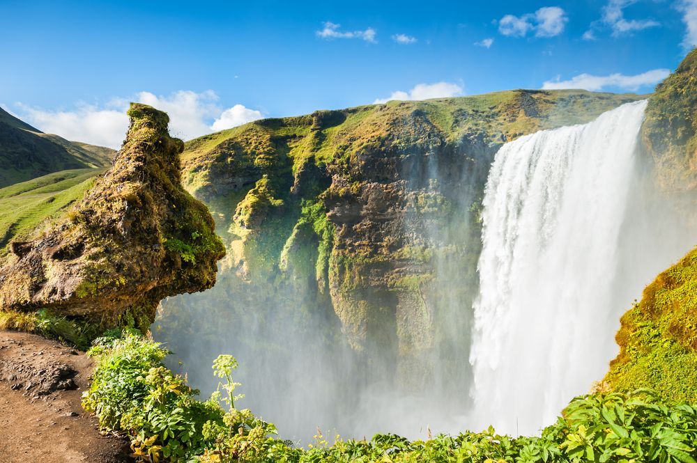 Waterfall plunging over green cliffs under a blue sky, with mist rising from the canyon below