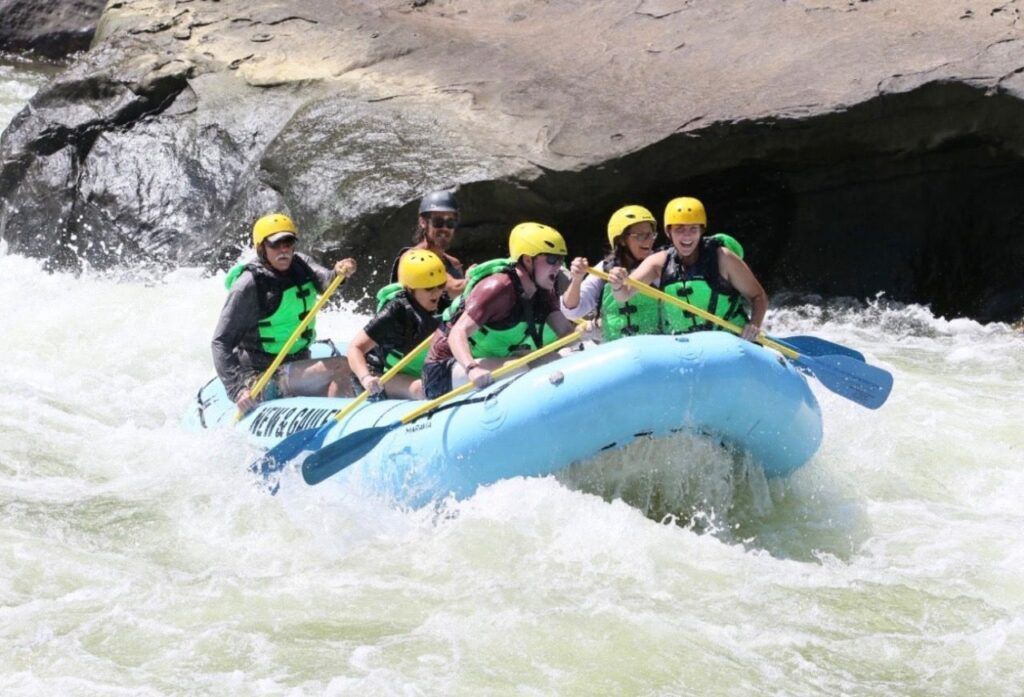 Rafting team in yellow helmets splashes through whitewater under a rocky cave opening
