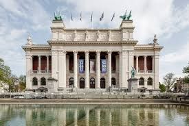 Grand neoclassical building with columns and statues reflected in a calm pool under a cloudy sky