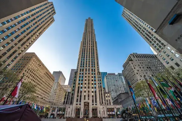View up at Rockefeller Center’s tall Art Deco tower, framed by surrounding skyscrapers and flags below