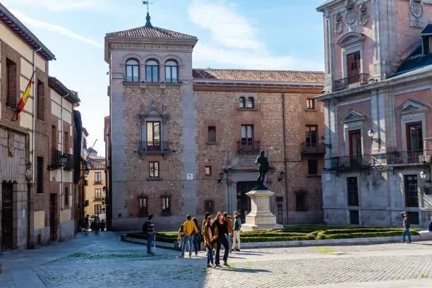 Historic stone plaza with a fountain and statue, people walking, and old buildings in the background