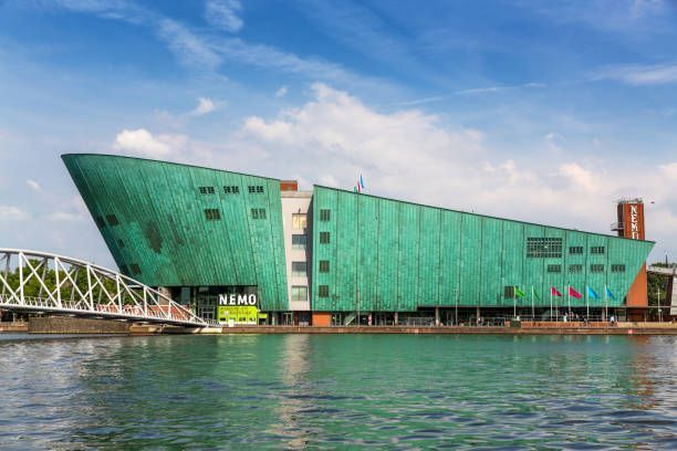 Green angular waterfront building with bridge and flags reflected in calm water under a cloudy sky