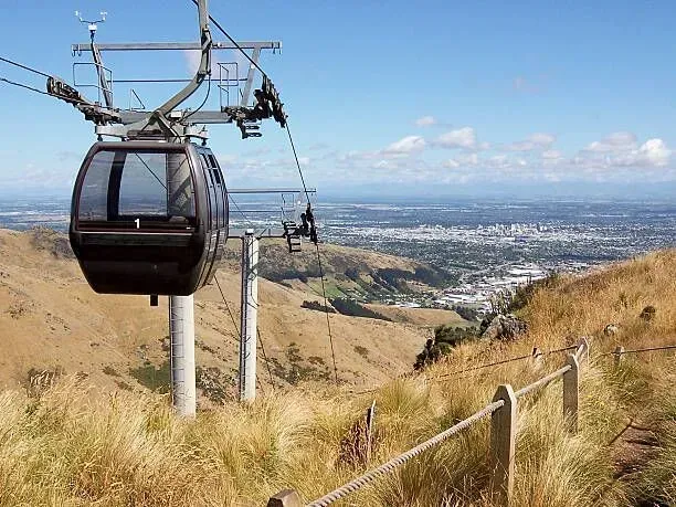 A black cable car glides over a dry hillside with a city skyline in the distance.