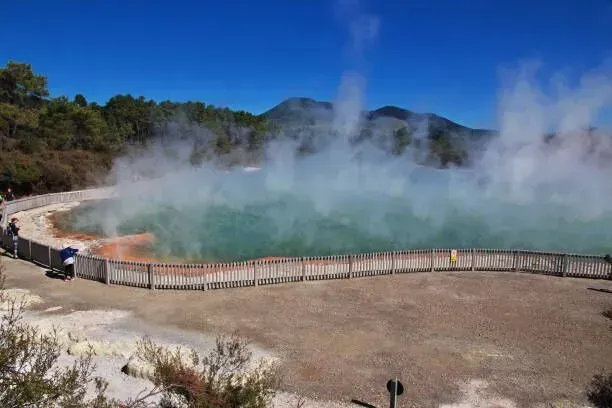 Steaming hot spring pool with turquoise water, fenced boardwalk, and mountains in the background