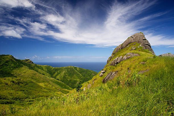 Green rolling hills and a rocky peak under a bright blue sky with wispy clouds