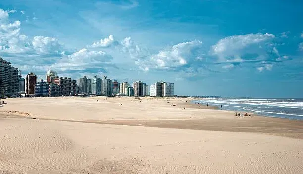 Wide sandy beach with calm ocean waves and a distant city skyline under a blue sky.