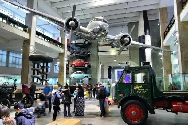 Museum exhibit with a silver airplane suspended above classic cars and a green truck, with visitors below