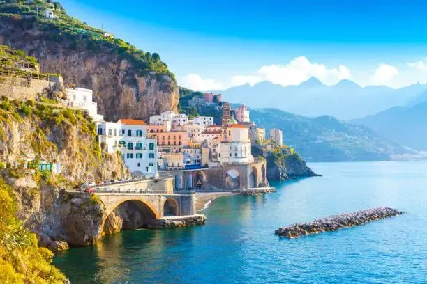 Colorful seaside village built into cliffs above turquoise water, with mountains in the distance.