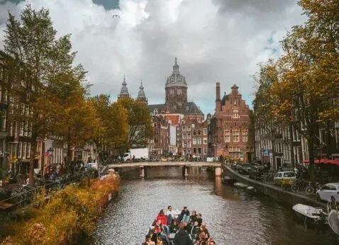 Canal with a boat, lined by autumn trees and historic buildings under a cloudy sky