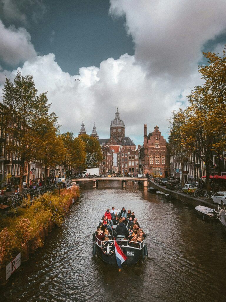 Canal boat gliding through a historic city canal under cloudy skies, framed by autumn trees and old buildings