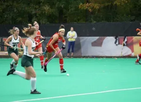 Field hockey players sprint after the ball on a green turf field during a match.