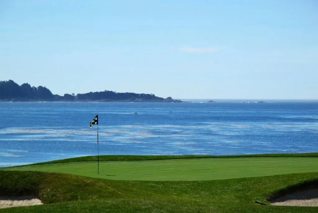 Golf green beside a blue ocean, with a flag near the water and distant shoreline in the background