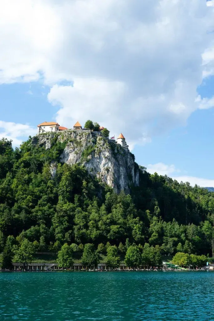 Cliffside castle overlooking a turquoise lake, framed by forest and cloudy blue sky