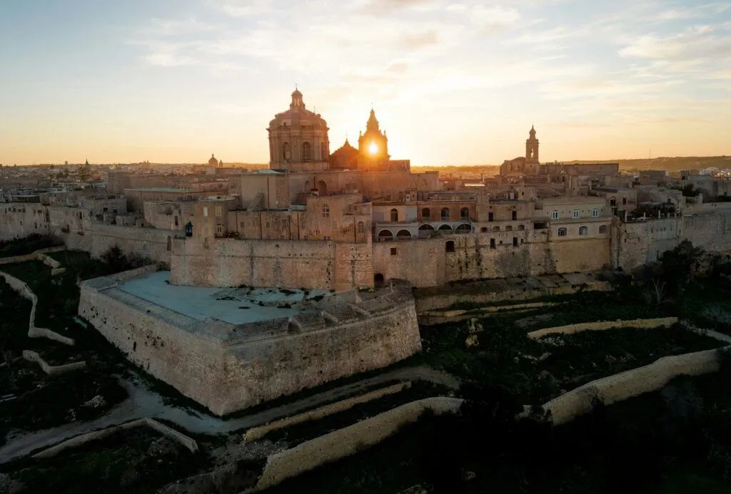 Sunset over a historic stone city with domed cathedral and fortress walls glowing in warm light