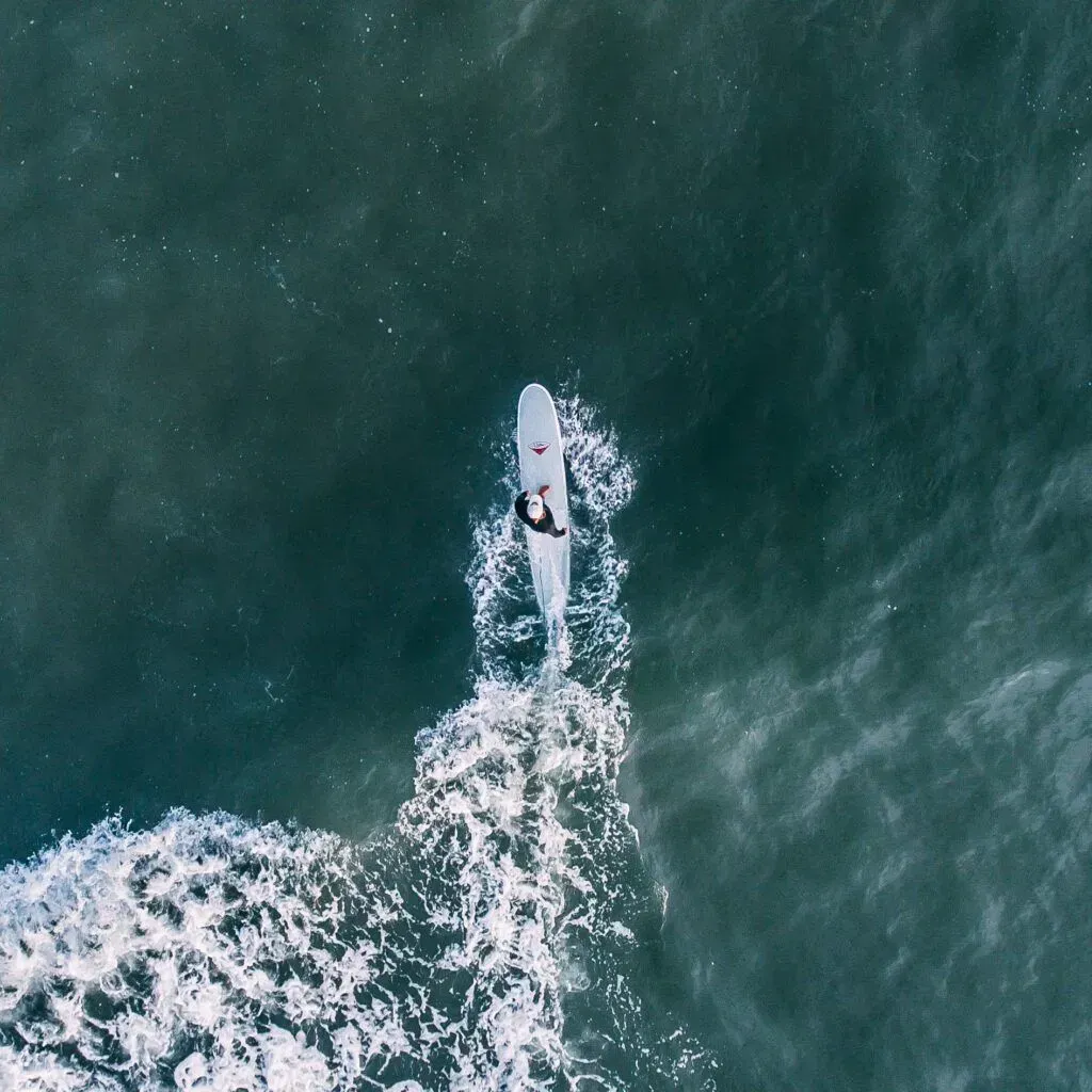 A lone surfer rides a white surfboard on dark green water, leaving a foamy wake.