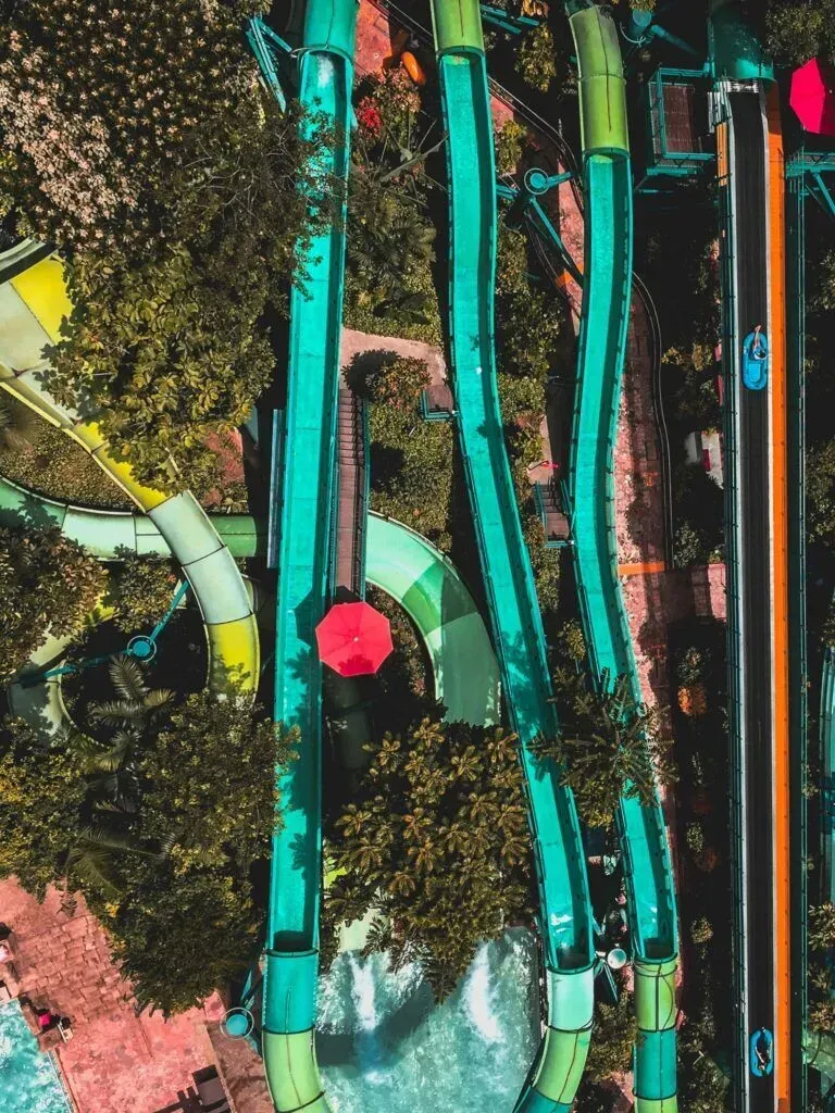 Abstract aerial view of colorful water slides winding through dense greenery, with a bright red umbrella below