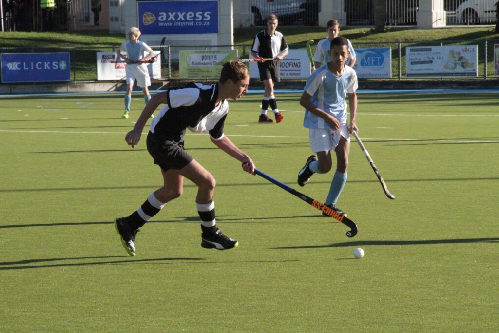 Field hockey players in black and white chase the ball on a green pitch during a match