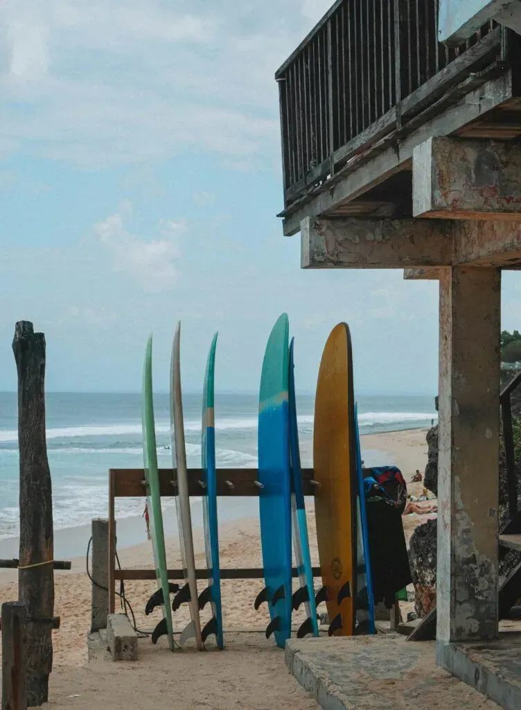 Surfboards lined up on a beach beside a weathered building, with ocean waves in the background.