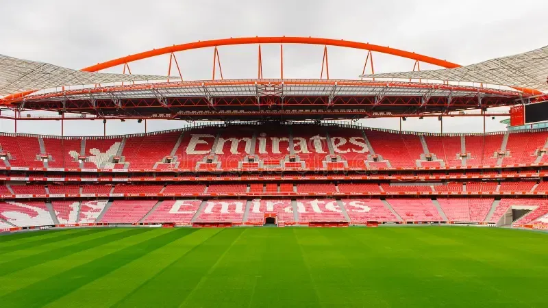Empty Emirates Stadium with red seats and green pitch under the roof arch.