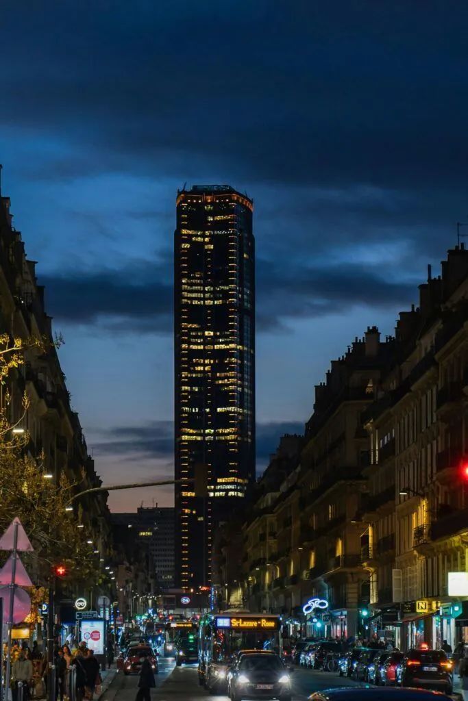 Night city street with traffic and lit buildings, centered on a tall illuminated tower under a dark blue sky
