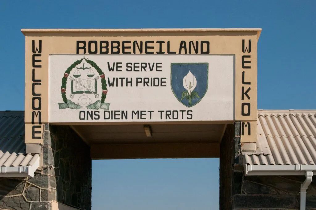 Robbeneiland entrance sign with “We serve with pride” and “Welcome” on a stone archway under a blue sky