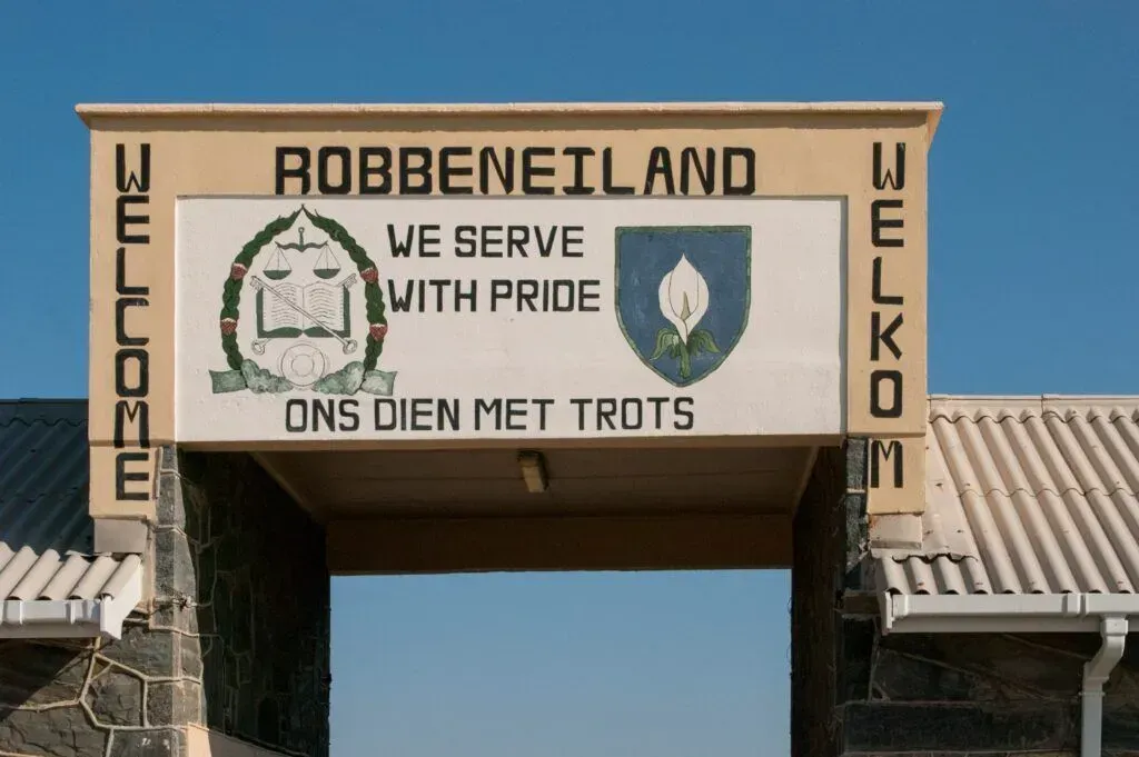 Entrance sign for Robben Island with “Welcome” and “Ons dien met trots” on a tan archway