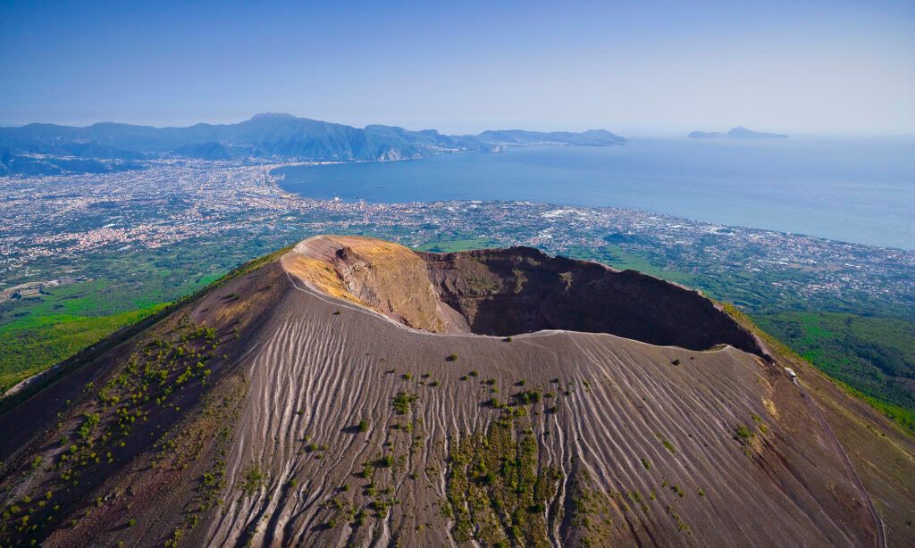 Aerial view of a volcanic crater with a rocky rim overlooking a green coastal landscape and city skyline