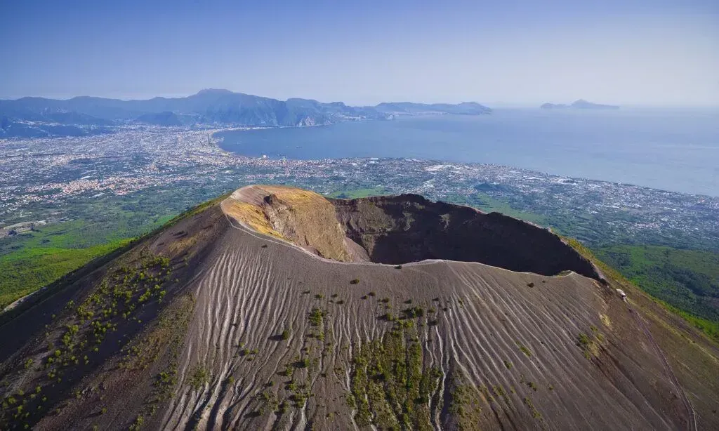 A volcanic crater with a steep rocky rim overlooking a green city and distant coastline