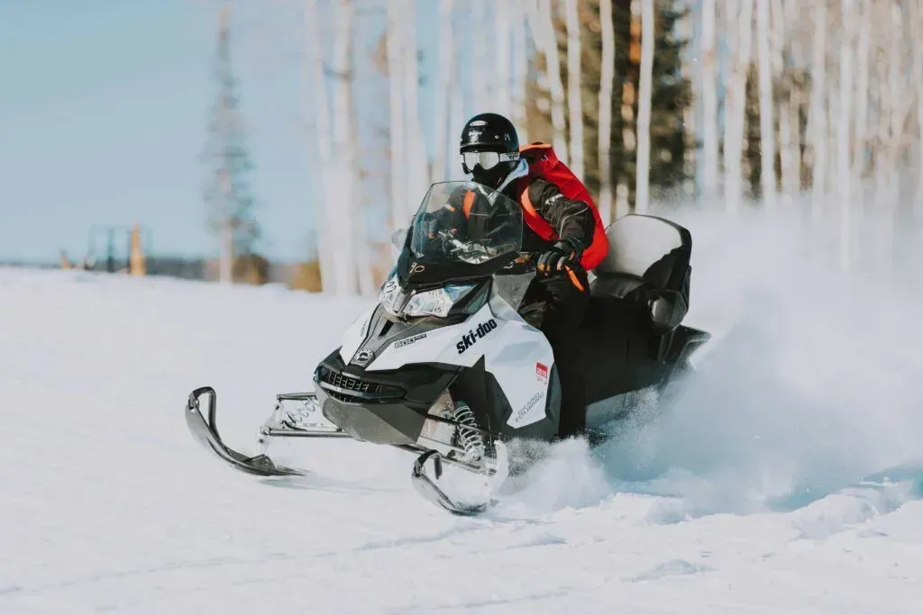 Snowmobiler in red and black racing through deep snow with a spray behind, in a snowy forest.