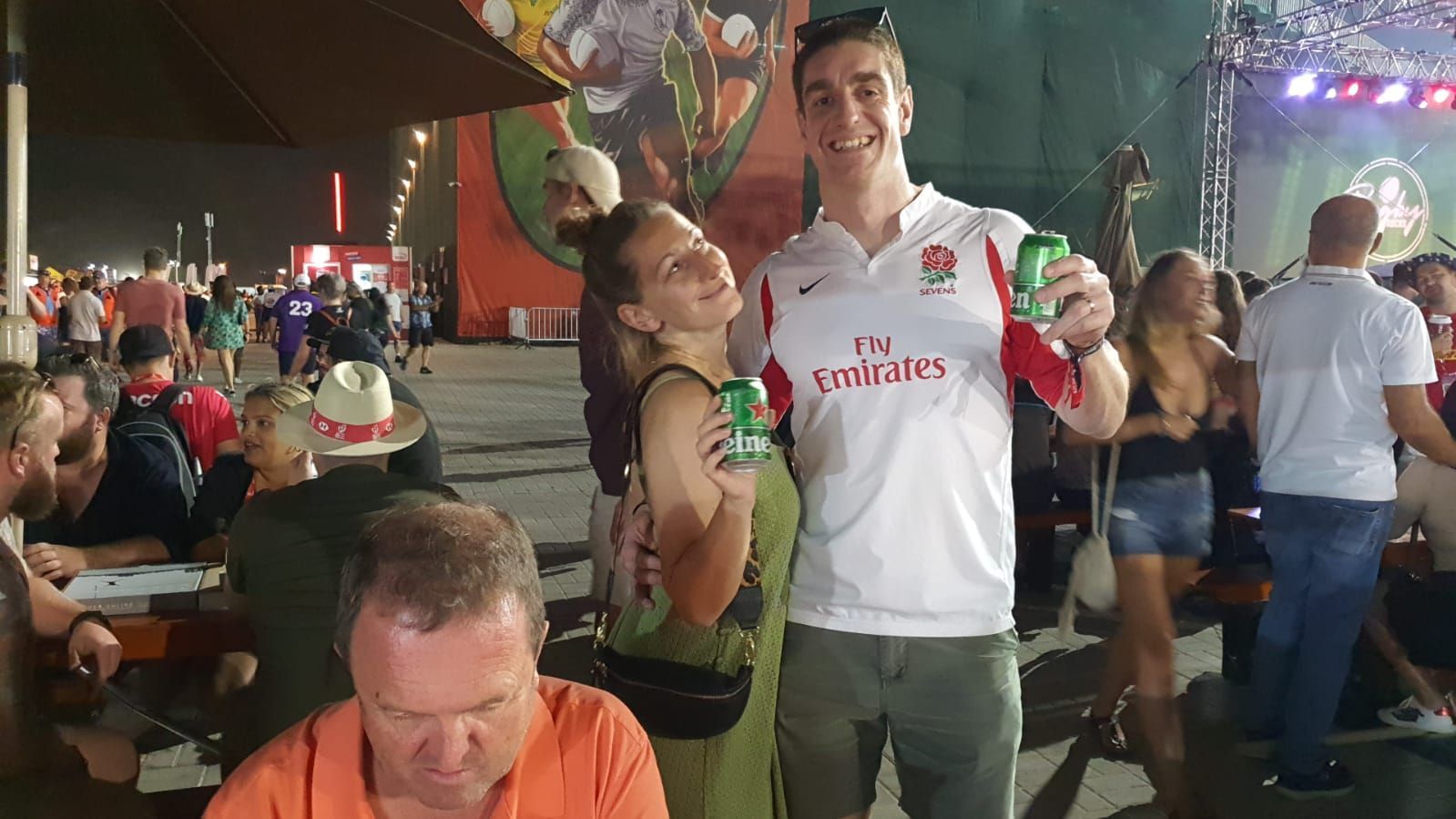 Crowded outdoor festival scene with people at tables; a smiling man in a white shirt holds a drink and poses with a woman.