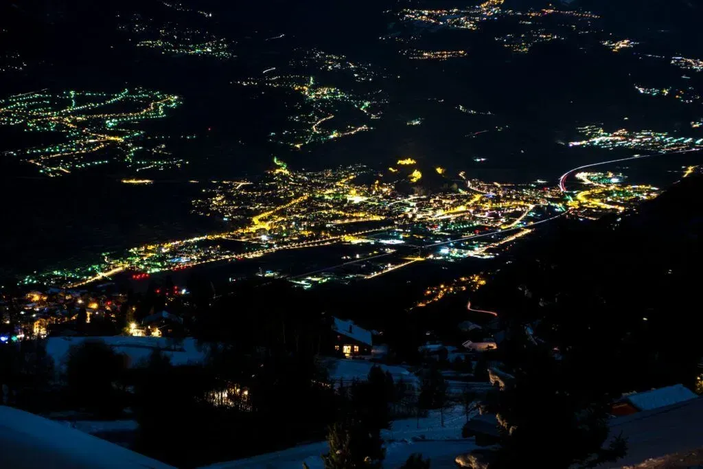 Nighttime view of a glowing valley town with scattered lights among dark hills and mountains