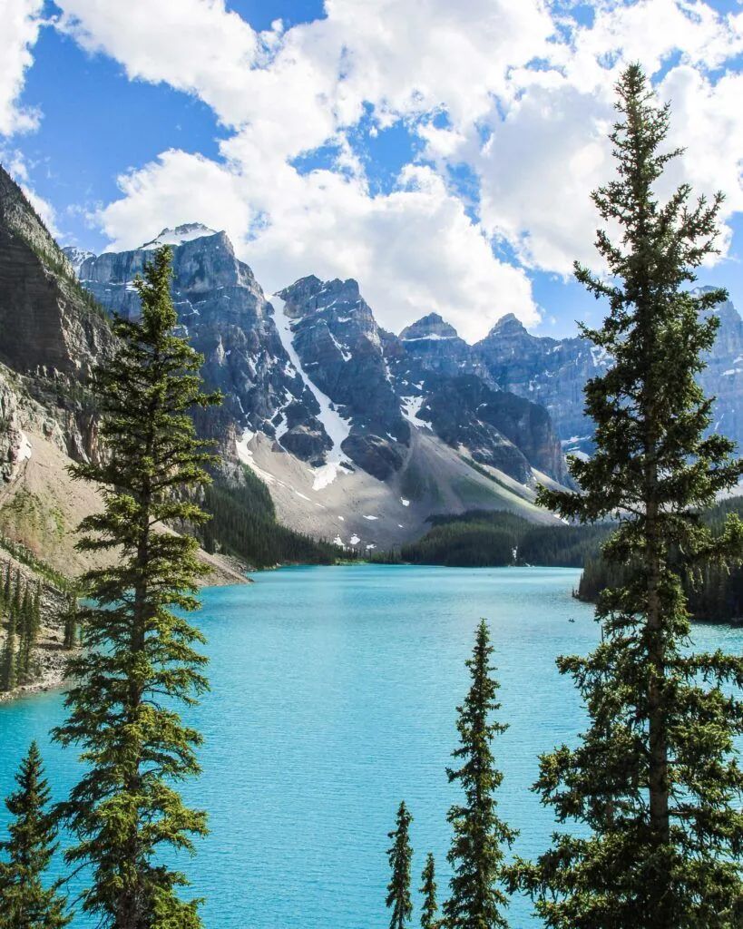 Turquoise alpine lake with pine trees and snow-dusted mountains under a partly cloudy sky