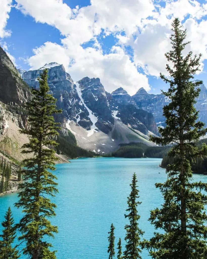 Turquoise alpine lake with pine trees and snow-dusted mountains under a cloudy blue sky