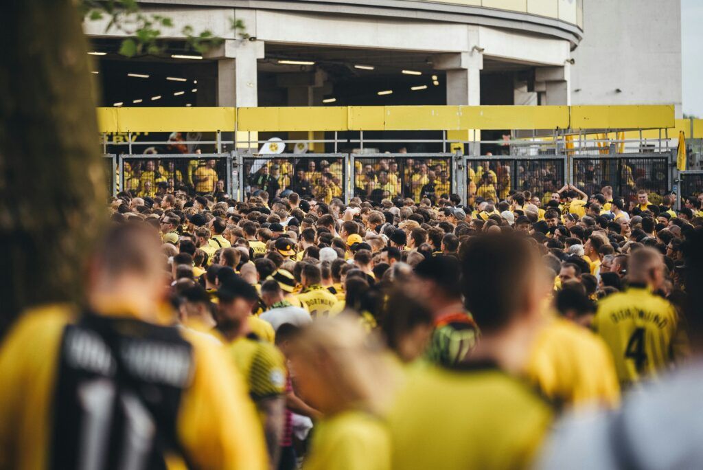Crowd of fans in yellow jerseys gathered outside a stadium entrance behind metal barriers