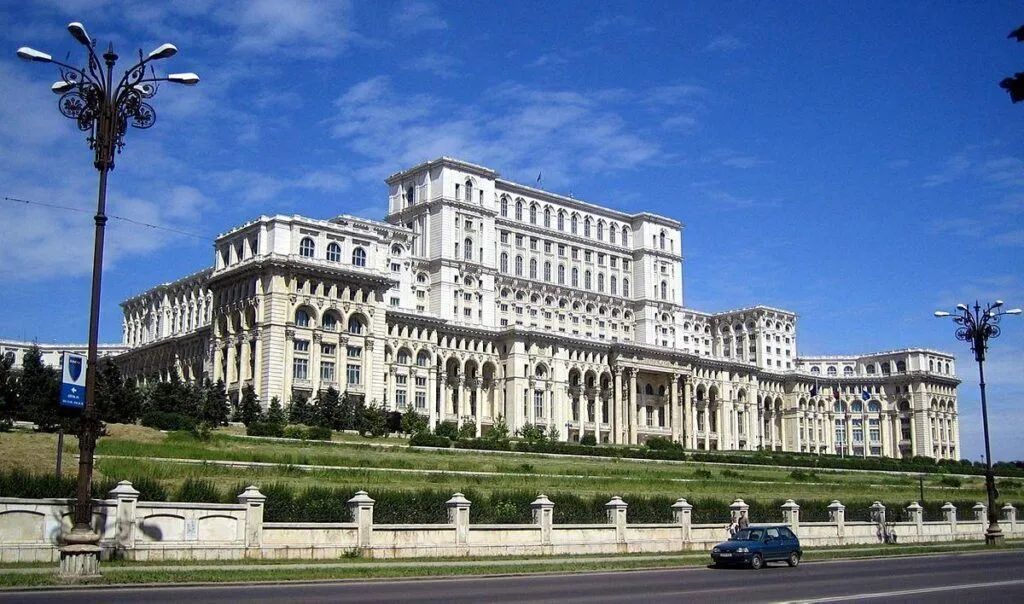 Large ornate government building with columns and a blue sky, viewed from a landscaped road