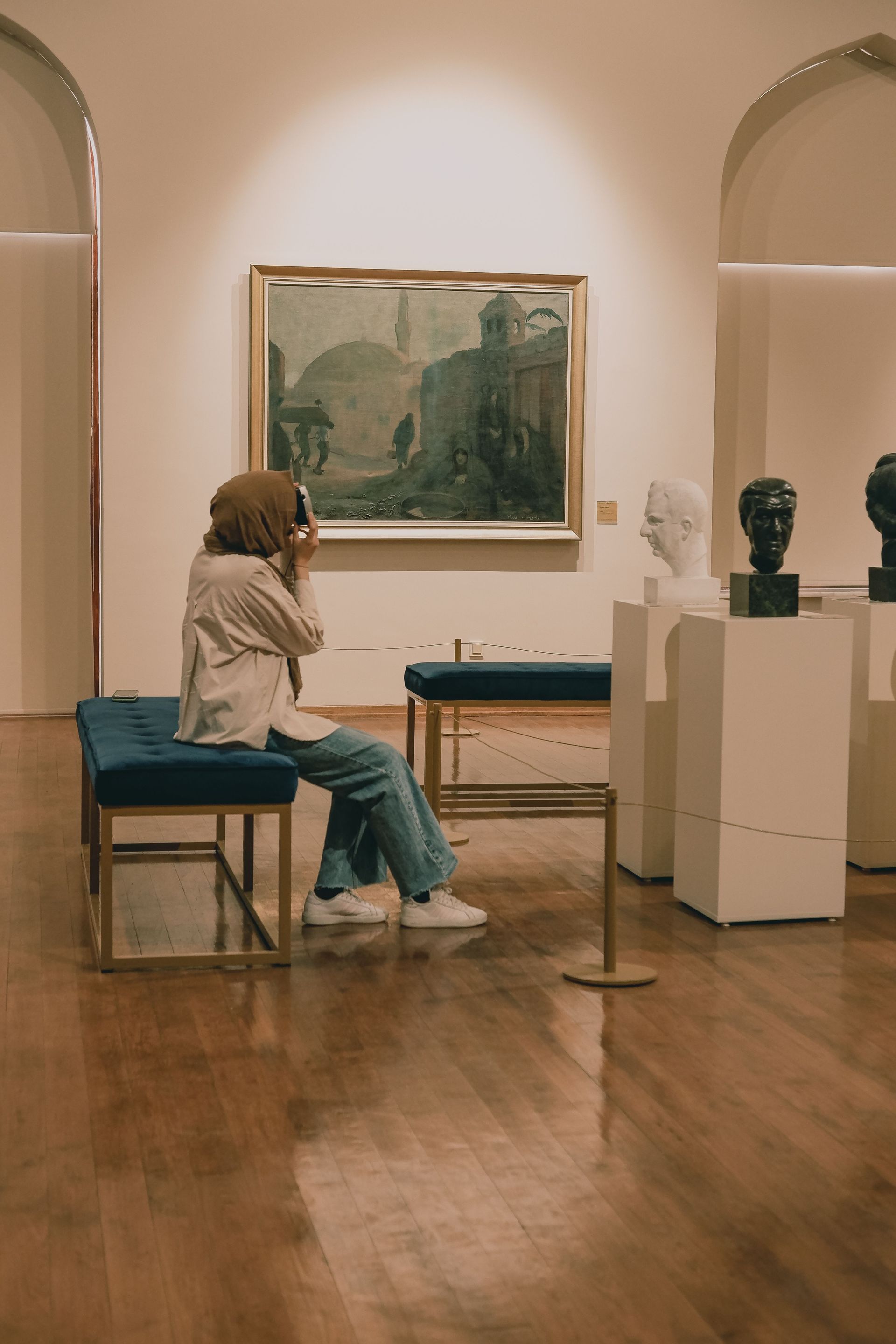 Person seated on a bench in an art gallery, looking at a landscape painting beside sculpture pedestals.