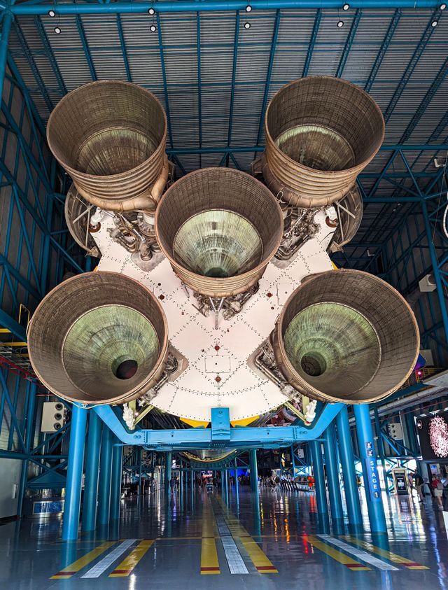 Five large rocket engine nozzles viewed from below in a blue industrial hangar or assembly bay