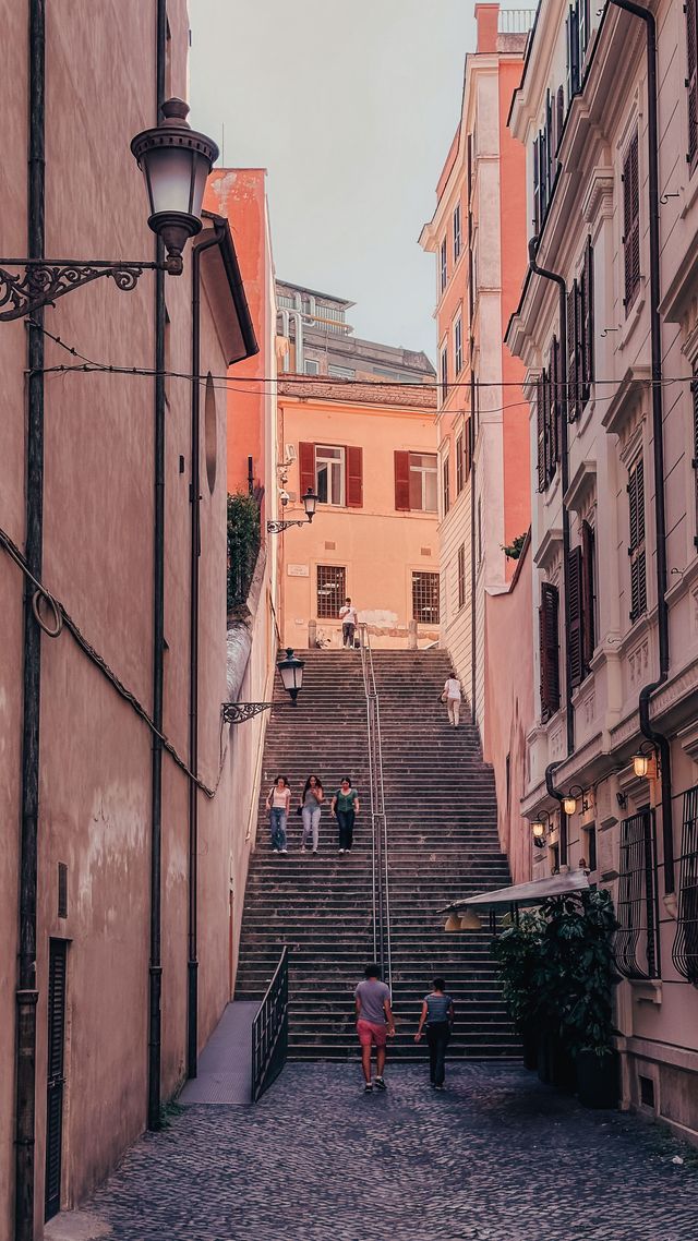 Narrow cobblestone alley with steep steps between tall orange buildings, two people walking up.