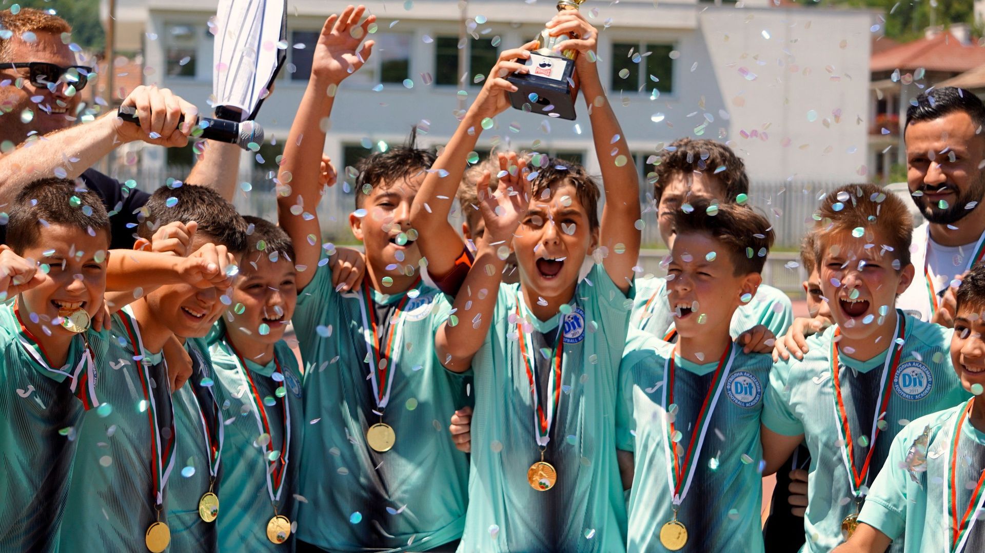 A group of athletes in teal jerseys celebrate with medals and raised trophies in a trophy-filled outdoor setting