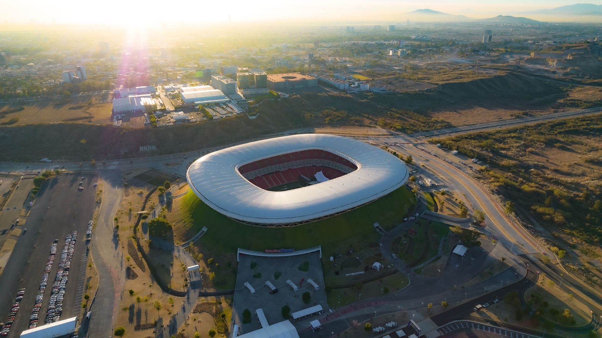Aerial view of a modern oval stadium with a white roof at sunset, surrounded by roads and city buildings.