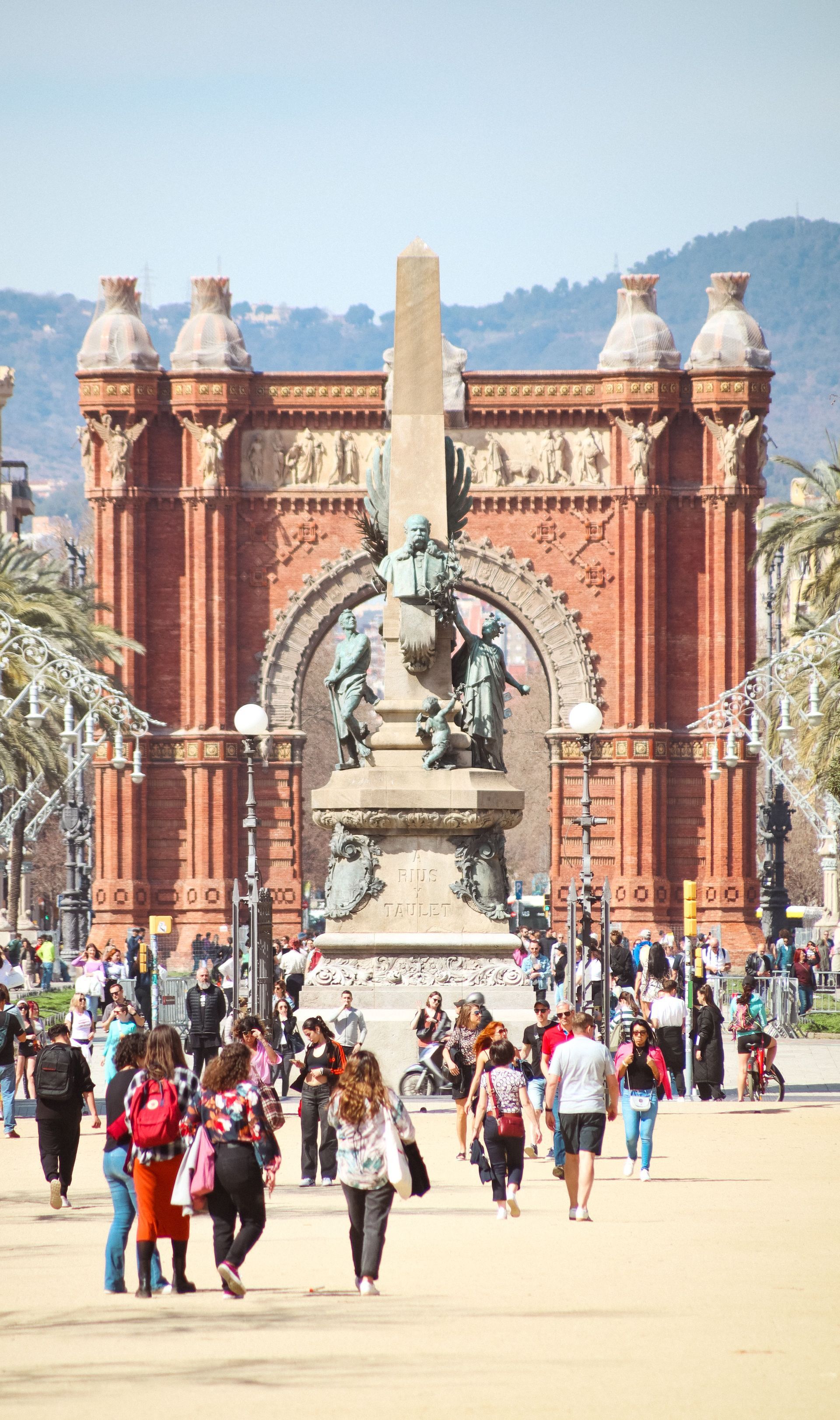 Crowded walkway leading to a red brick historic arch and statue in front of a museum-like building