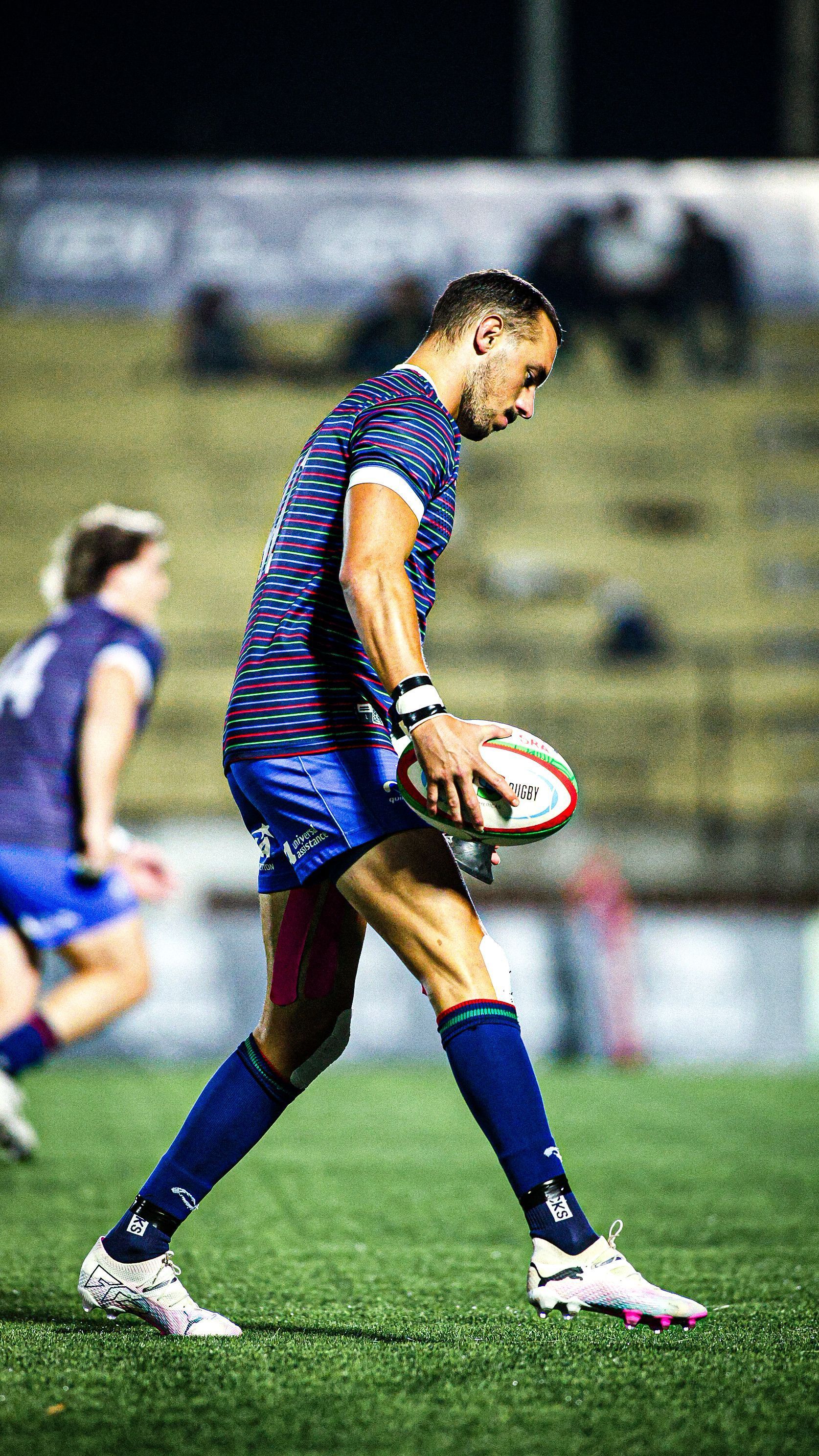 Rugby player in blue and red uniform runs on a field holding the ball at night