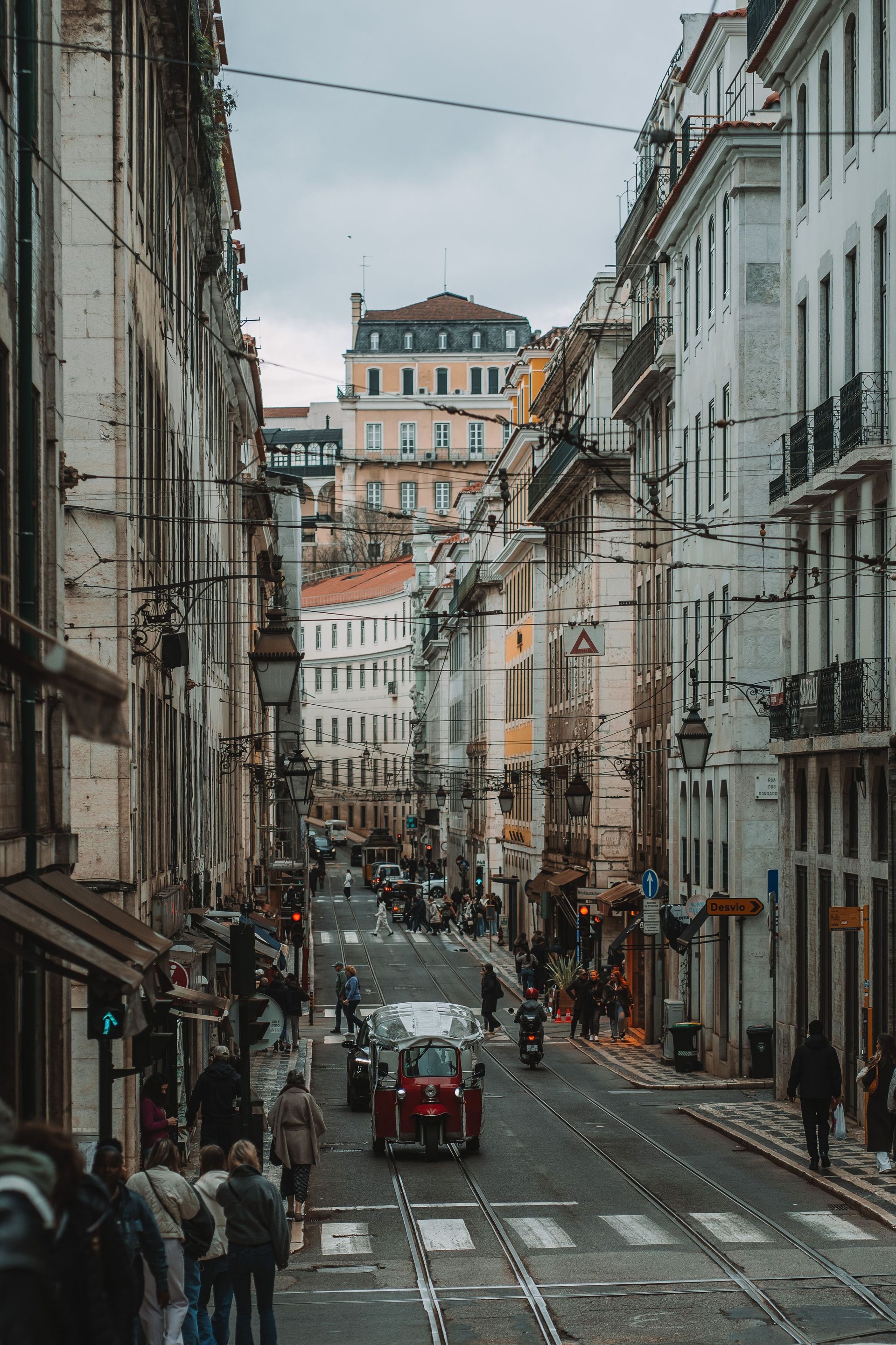 Narrow European street with tall buildings, pedestrians, and a red vehicle under overcast skies