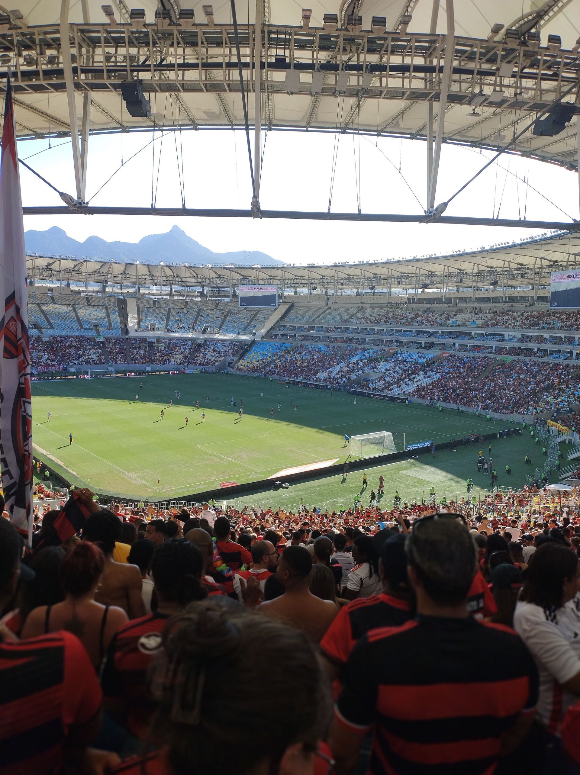 Crowded soccer stadium with fans in red-and-black shirts, viewed from the stands under a roof.