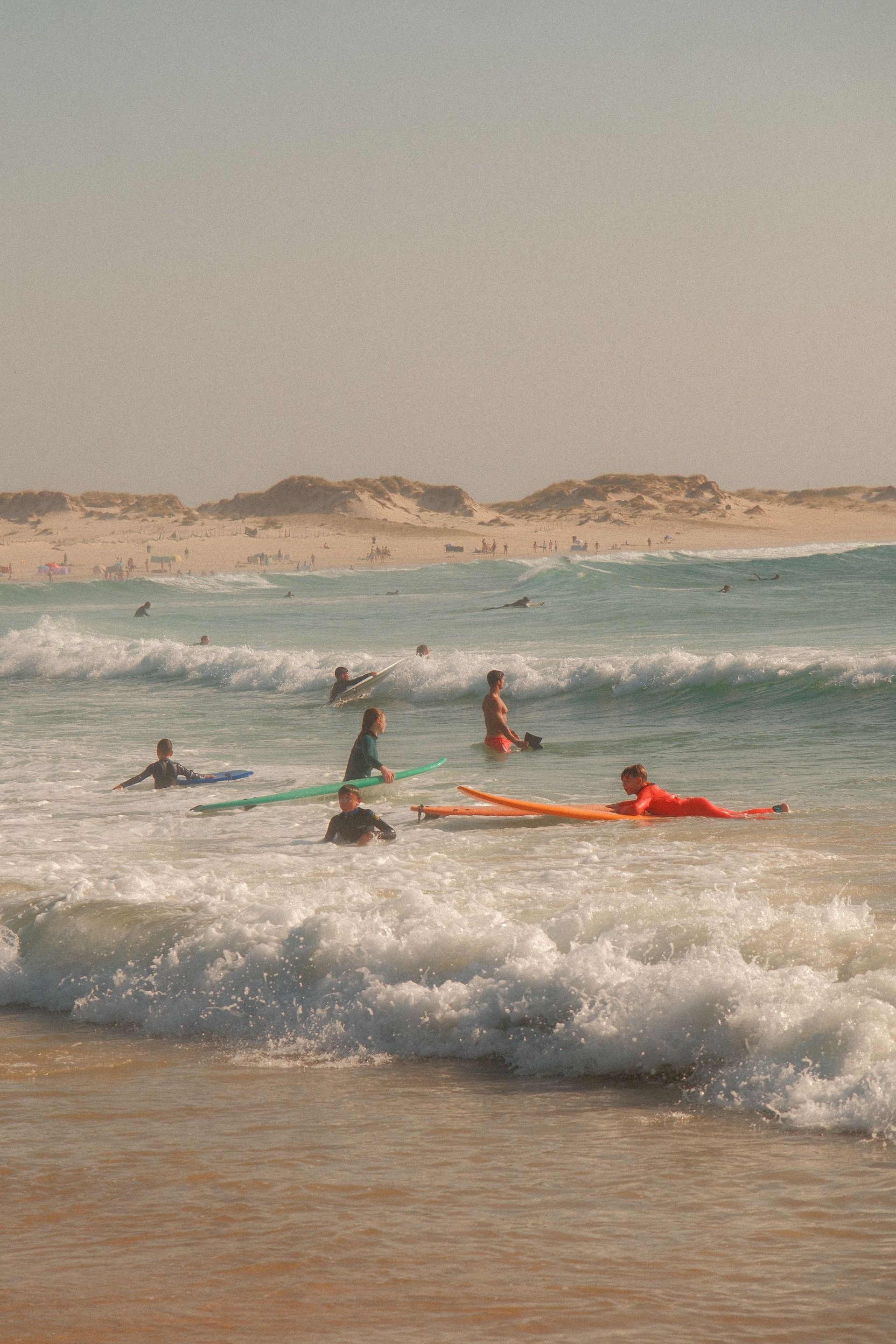Surfers paddling on waves at a sandy beach with distant dunes under a hazy sky